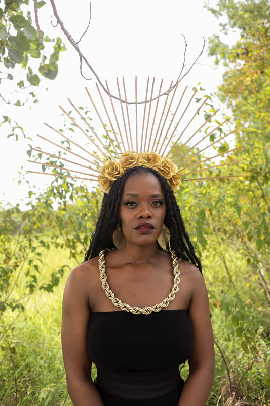 Ebony Stewart, a thin dark-skinned Black woman, stares supremely at the viewer. She is wearing a large spiky gold flower crown, oval shaped earrings, a thick gold dookie rope, and a black tube top. She is standing in a field.